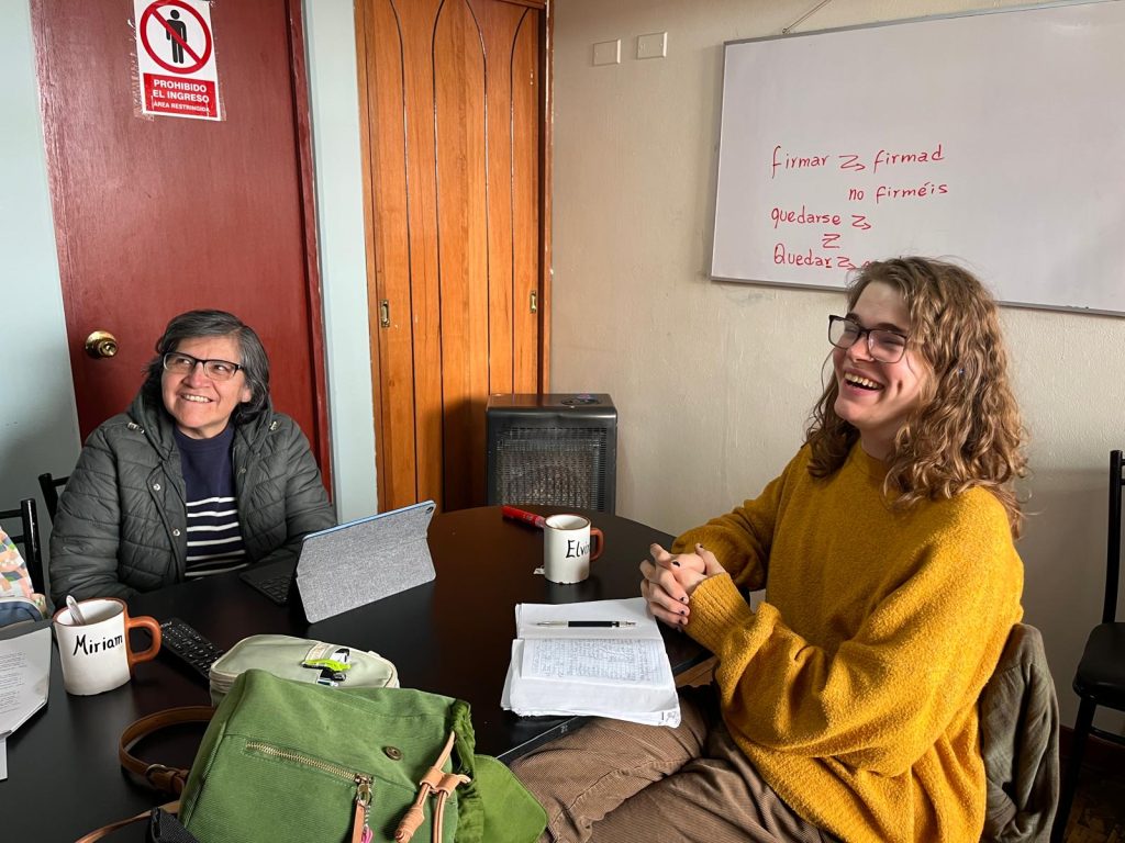 Local Cusqueña Spanish teacher and foreign American student smiling together while sitting in a naturally lit classroom with Spanish lesson behind them on whiteboard.